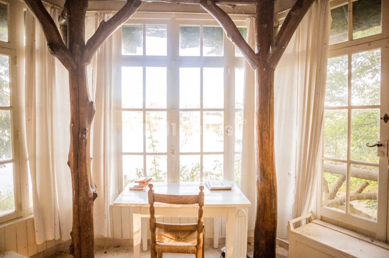 Bureau en bois clair avec une chaise, encadré par des troncs d'arbres, devant une grande baie vitrée lumineuse.