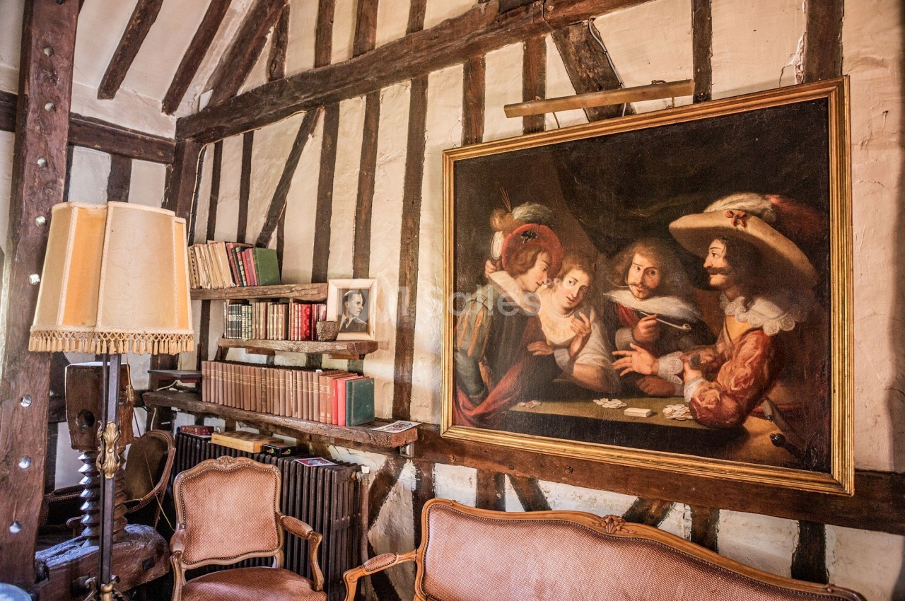 Intérieur rustique avec poutres apparentes, bibliothèque, fauteuils anciens et tableau représentant une scène de cartes.