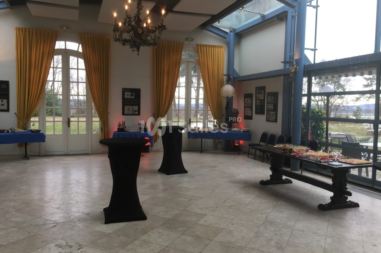Salle lumineuse avec grandes fenêtres, rideaux jaunes, tables hautes noires et buffet disposé sur une table en bois.