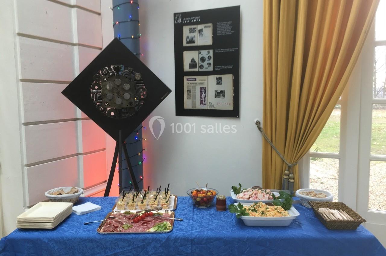 Buffet avec nappe bleue, proposant charcuterie, salades, brochettes et pains, dans une salle décorée de rideaux jaunes.