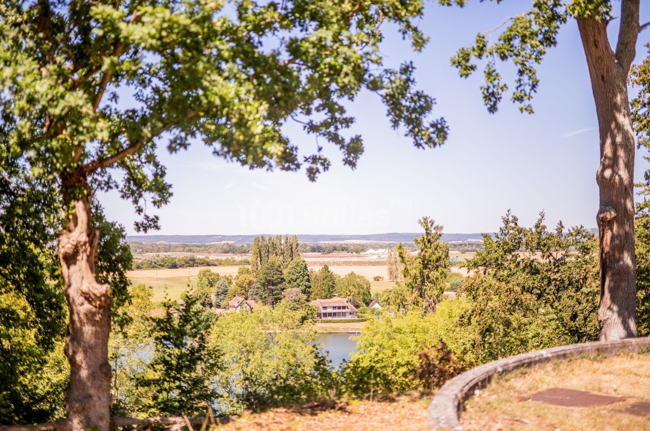 Vue sur un paysage rural avec des arbres encadrant une rivière, des maisons et des champs à l'arrière-plan.