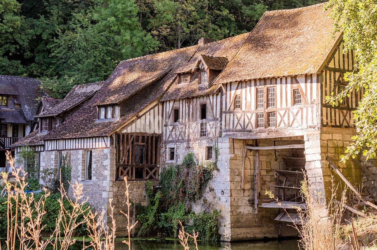 Maisons à colombages anciennes avec toits en tuiles, bordant une rivière et entourées de végétation dense.
