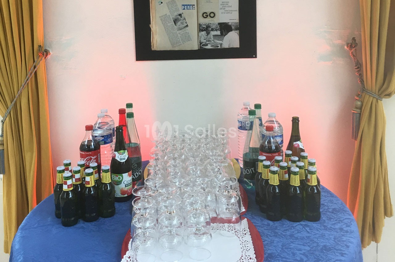 Table avec des bouteilles de boissons variées, verres vides alignés au centre et rideaux jaunes en arrière-plan.