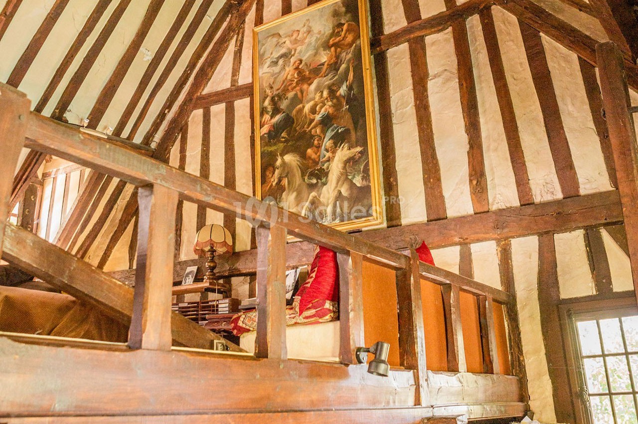 Intérieur d'une pièce avec poutres apparentes, mezzanine en bois, tableau accroché et lumière naturelle entrant par une…