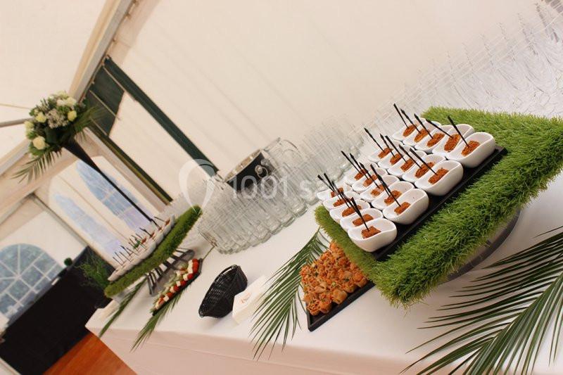 Buffet avec verrines et amuse-bouches disposés sur des plateaux décorés de gazon artificiel dans une salle lumineuse.