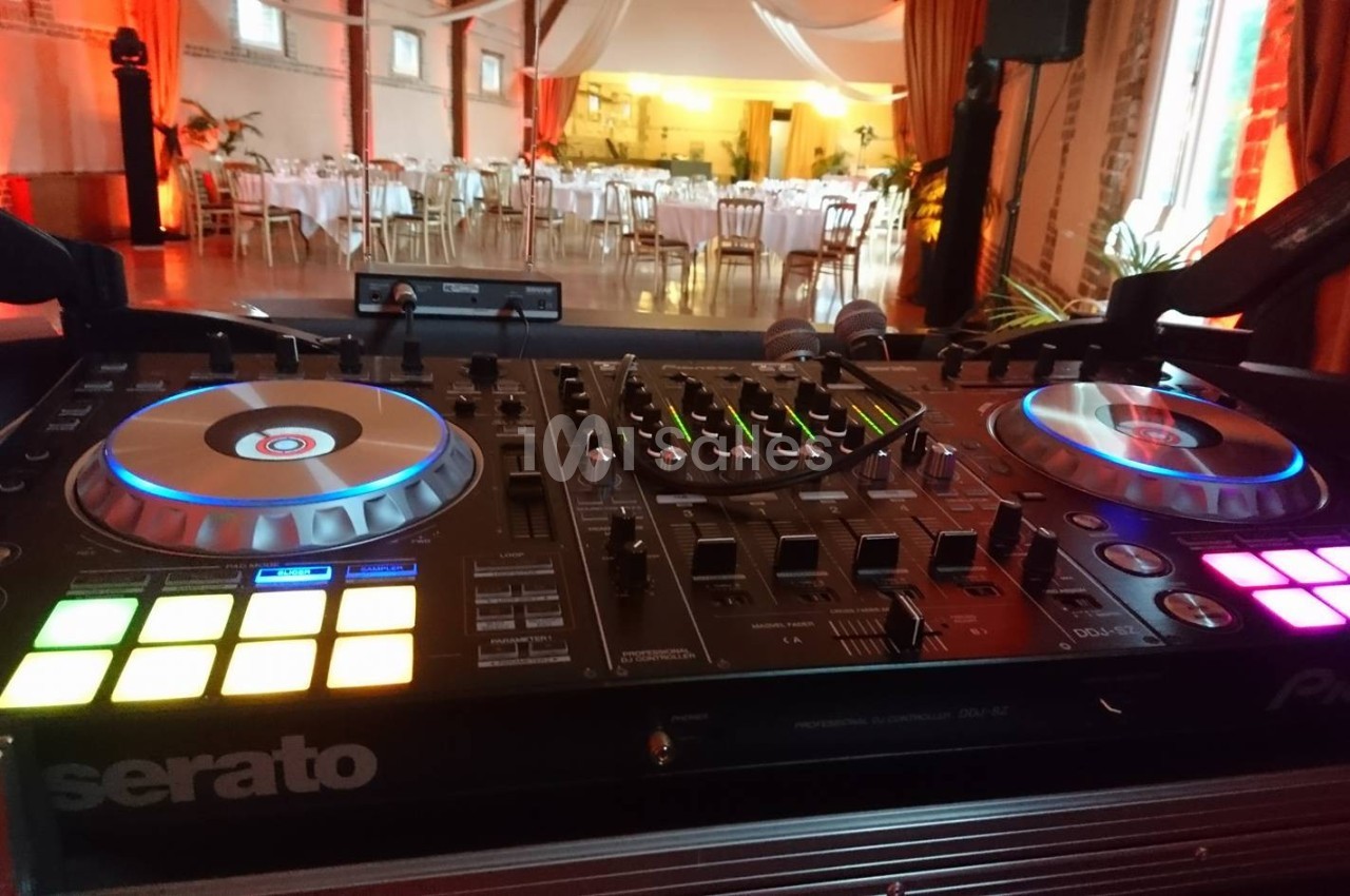 Table de mixage DJ avec platines lumineuses en premier plan, salle de réception décorée et tables dressées en arrière-plan.