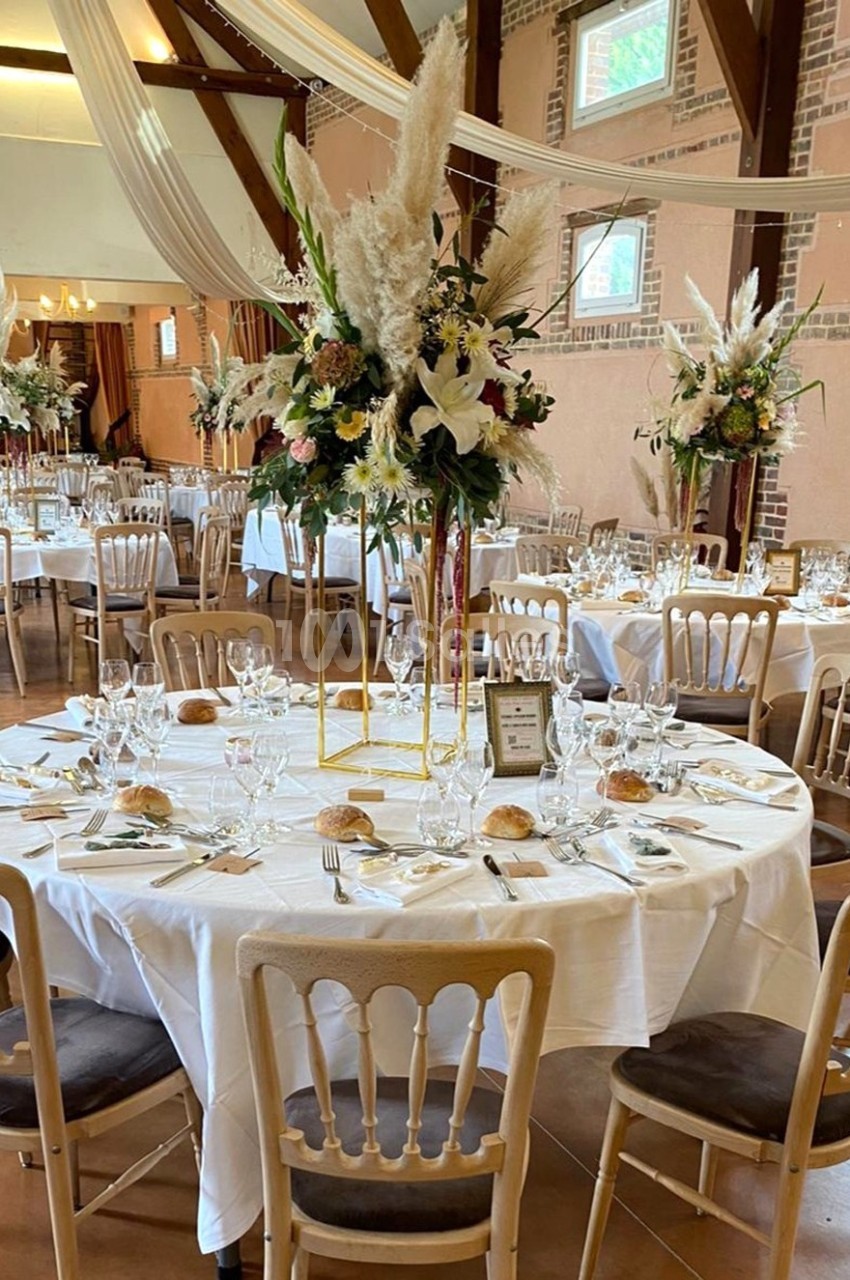 Salle de réception décorée avec des tables rondes dressées, ornées de centres de table floraux et de chaises en bois clair.