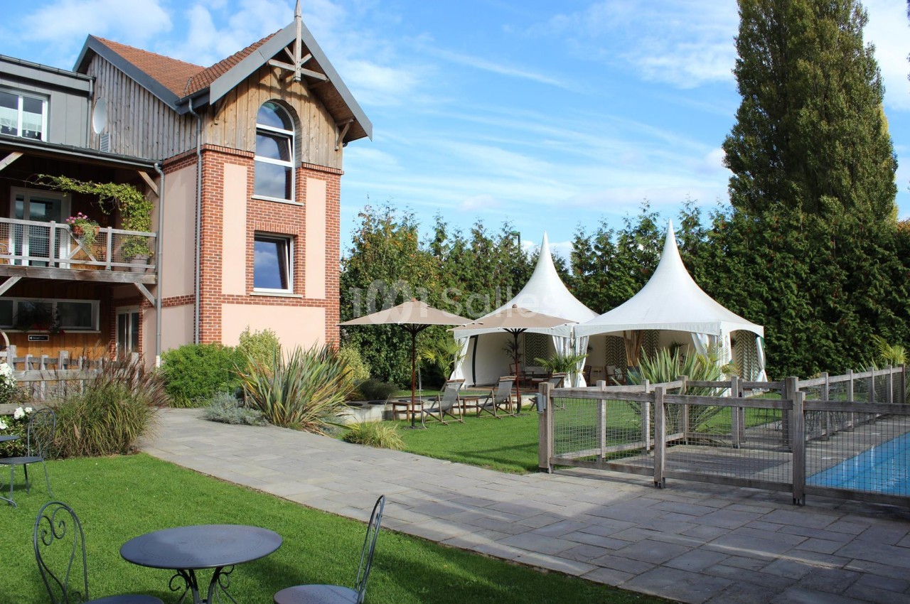 Maison en briques avec jardin, terrasse, tentes blanches et piscine clôturée sous un ciel bleu.