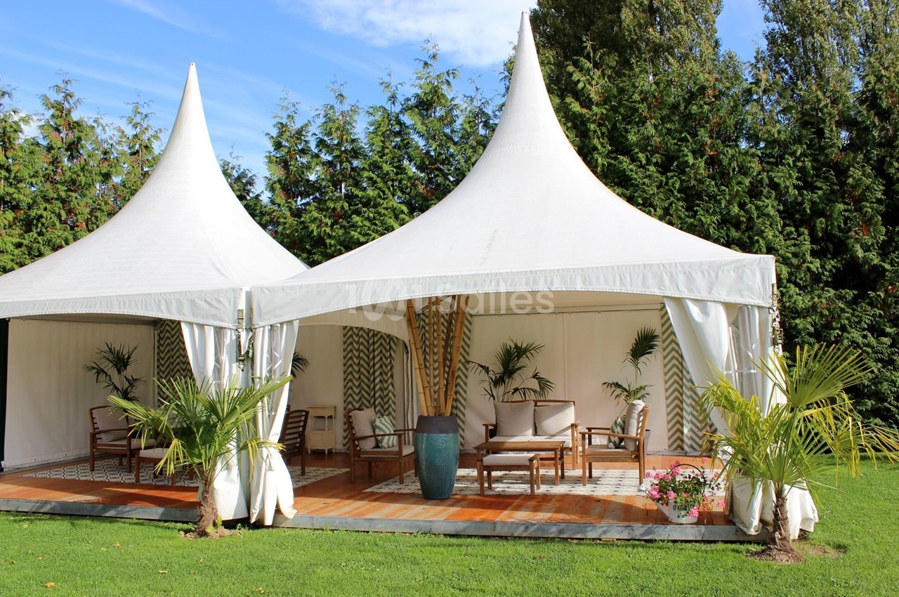 Deux tentes blanches avec mobilier en bois, décorées de plantes, installées sur une pelouse devant des arbres.