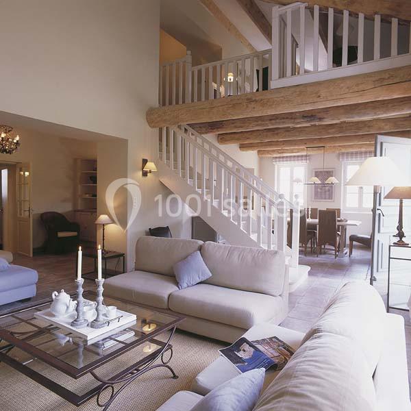 Location salle Sorgues (Vaucluse) - Château La Tour Vaucros #22 Salon lumineux avec poutres apparentes, canapé beige, table basse en verre et escalier menant à une mezzanine.