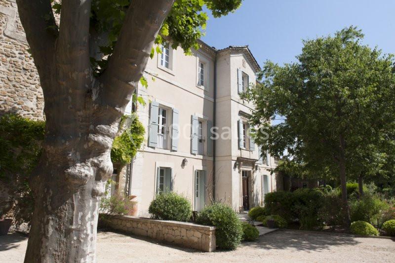 Façade d'une maison en pierre claire avec volets bleus, entourée d'arbres et de végétation sous un ciel dégagé.