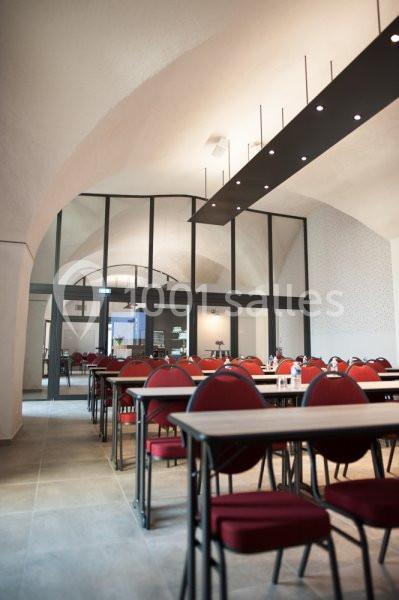 Location salle Sorgues (Vaucluse) - Château La Tour Vaucros #15 Salle lumineuse avec des tables et des chaises rouges alignées, sous un plafond voûté et éclairée par des suspensions…