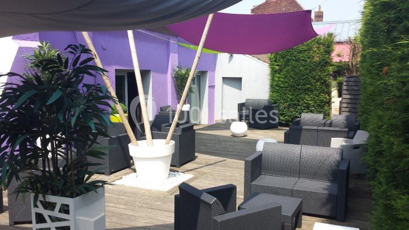 Terrasse aménagée avec fauteuils en rotin, plantes en pot et voiles d'ombrage colorées, entourée de haies.