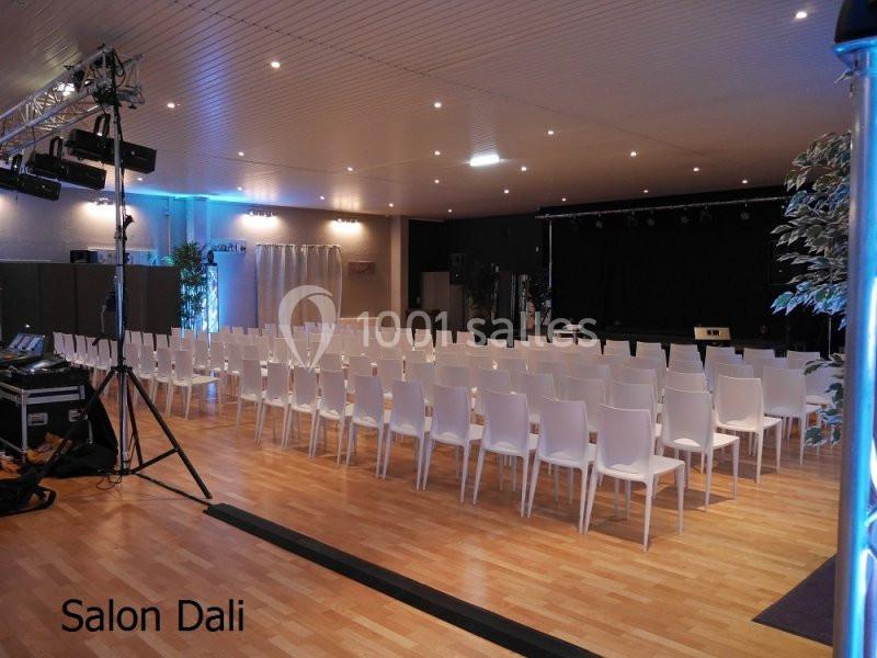 Salle de conférence avec des rangées de chaises blanches, une scène au fond et un éclairage tamisé.