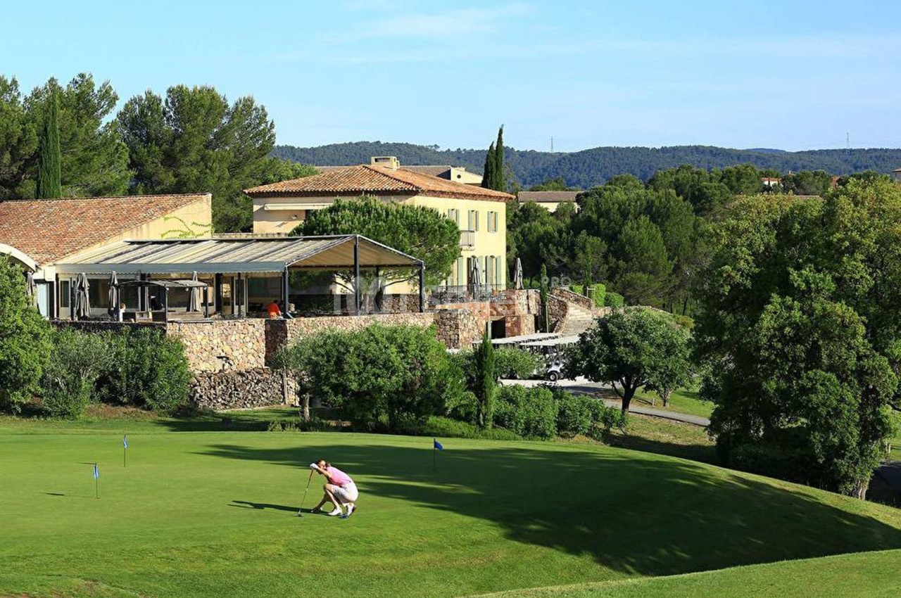 Un golfeur ajuste son coup sur un green entouré de végétation, avec des bâtiments en arrière-plan.