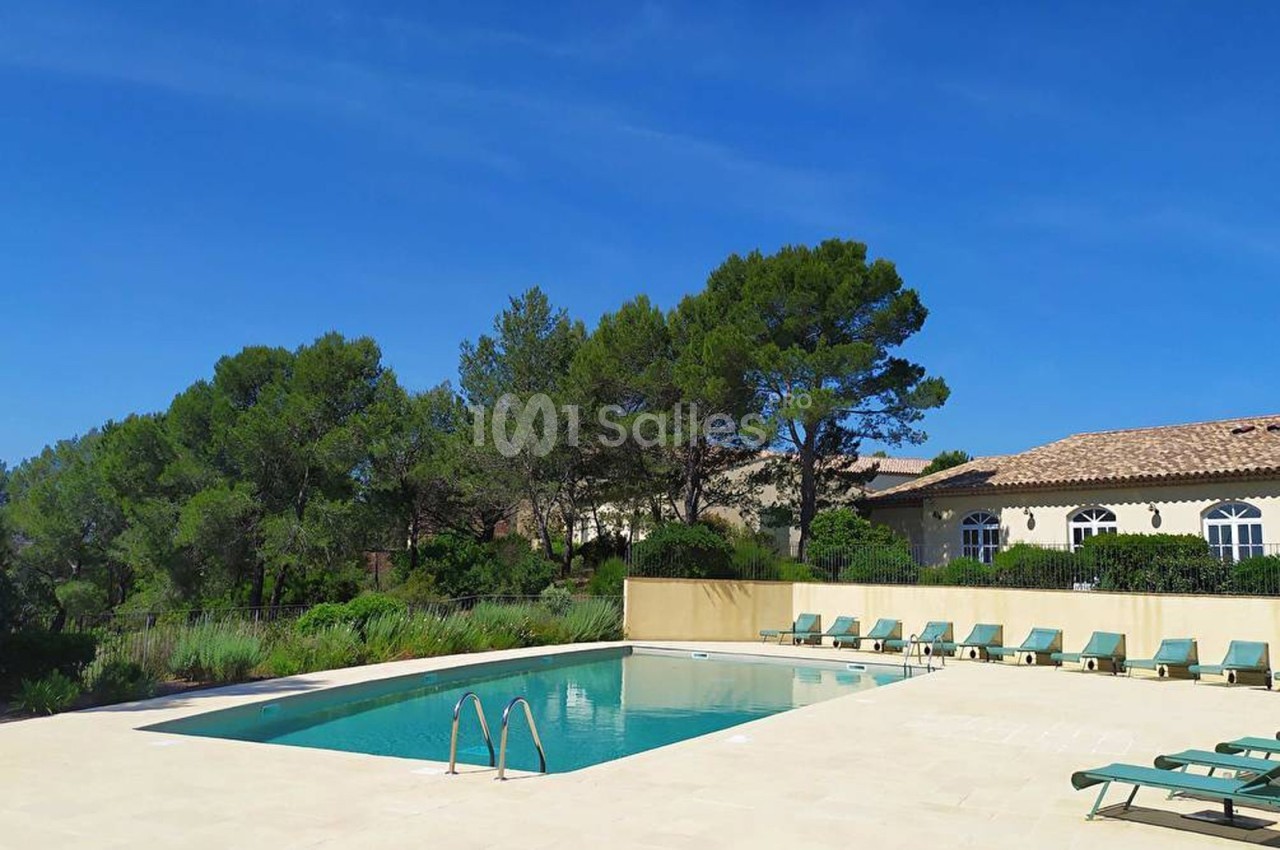 Piscine extérieure entourée de chaises longues, bordée de végétation et d'un bâtiment avec toit en tuiles sous un ciel bleu.
