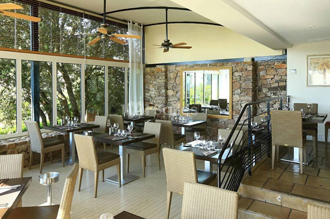 Salle de restaurant lumineuse avec tables dressées, chaises en osier, murs en pierre et grandes baies vitrées donnant sur…