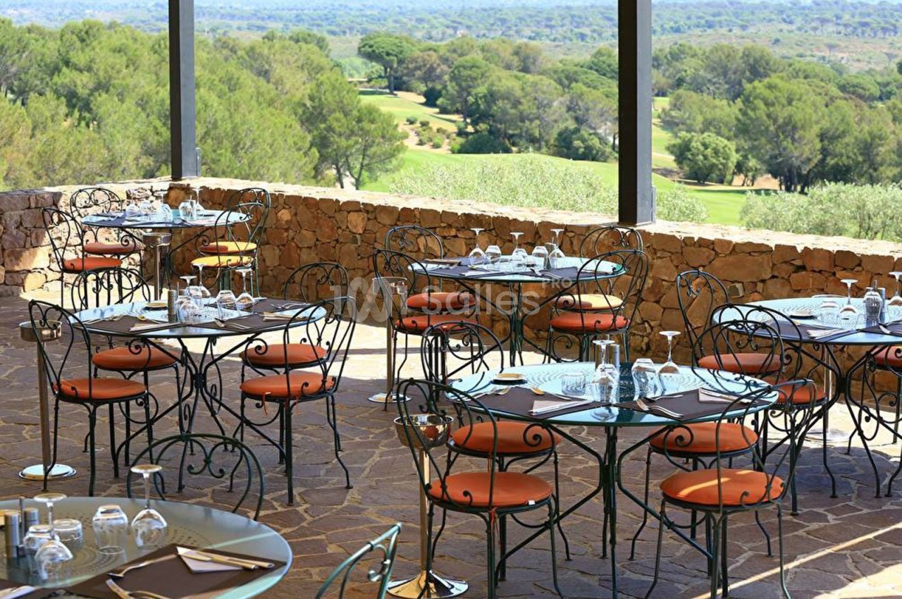 Terrasse d'un restaurant avec des tables en fer forgé, vue sur un paysage arboré et un terrain de golf.