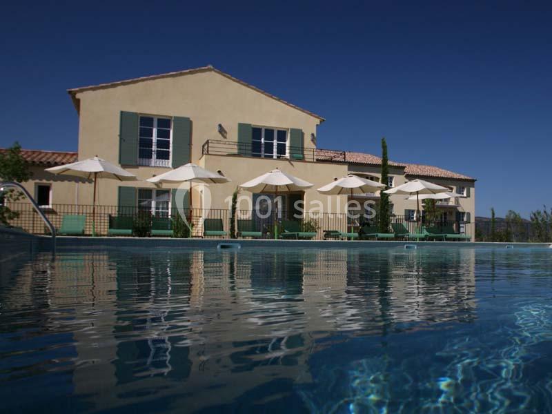 Maison provençale avec volets verts, terrasse et piscine, entourée de chaises longues et parasols sous un ciel bleu.
