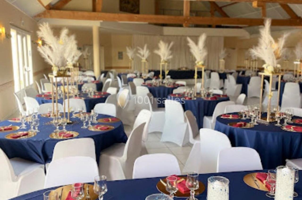 Salle de réception décorée avec des tables rondes nappées de bleu, ornées de centres de table et de vaisselle élégante.
