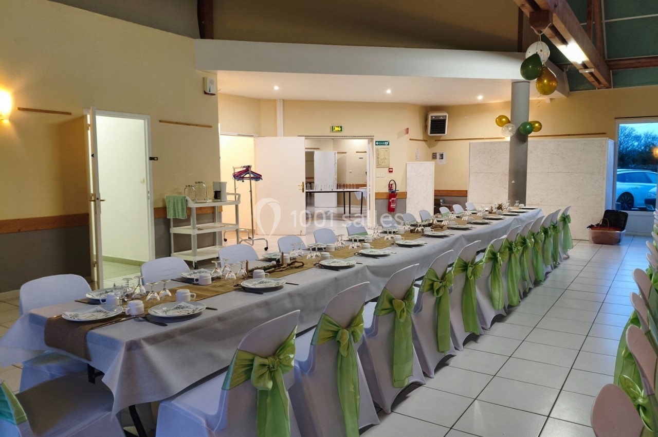 Salle décorée avec une longue table dressée pour un repas, chaises ornées de nœuds verts et ballons suspendus.