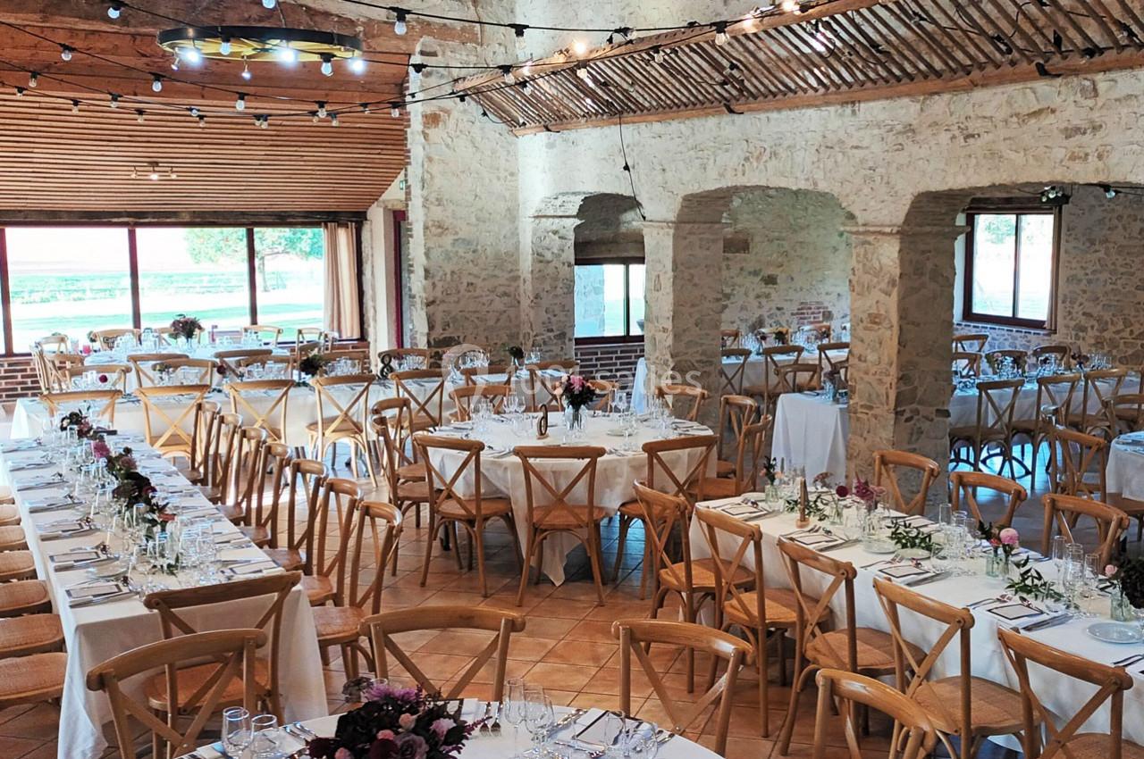 Salle de réception en pierre avec tables décorées, chaises en bois et guirlandes lumineuses suspendues.