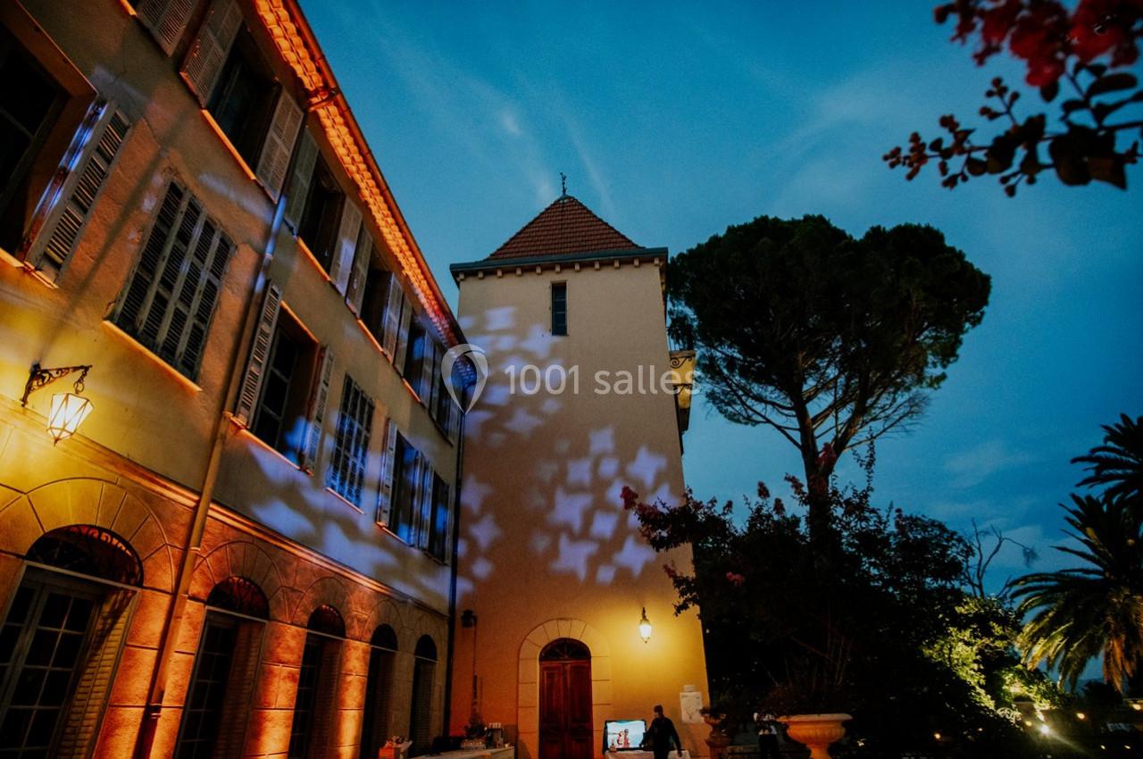 Location salle Le Cannet-des-Maures (Var) - Château Colbert Cannet #13 Façade d'un bâtiment éclairé par des lumières colorées au crépuscule, entouré d'arbres et de végétation.