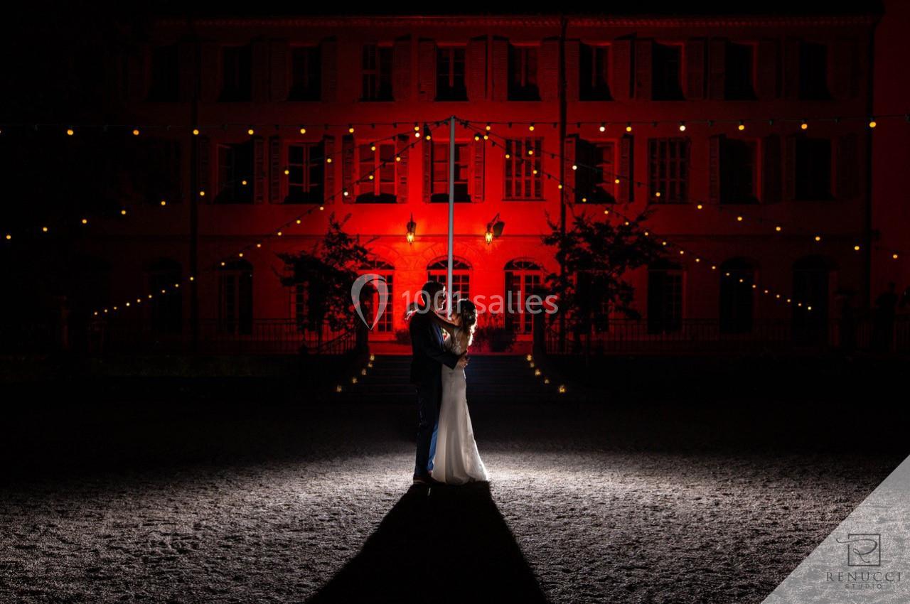 Location salle Le Cannet-des-Maures (Var) - Château Colbert Cannet #17 Un couple danse sous des guirlandes lumineuses devant un bâtiment éclairé en rouge, de nuit.