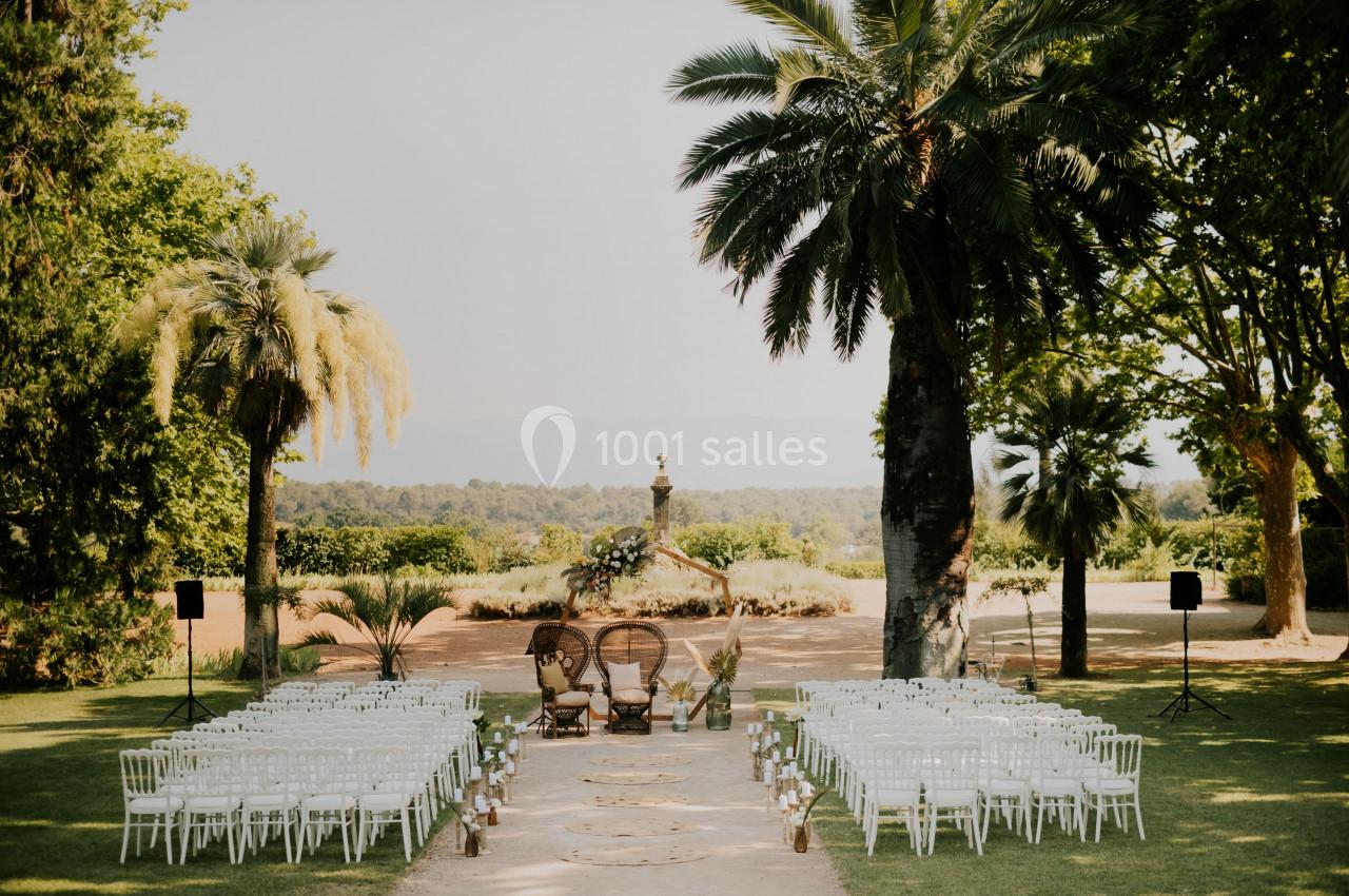 Location salle Le Cannet-des-Maures (Var) - Château Colbert Cannet #5 Allée centrale bordée de chaises blanches menant à deux fauteuils en osier, dans un jardin avec palmiers et fontaine.