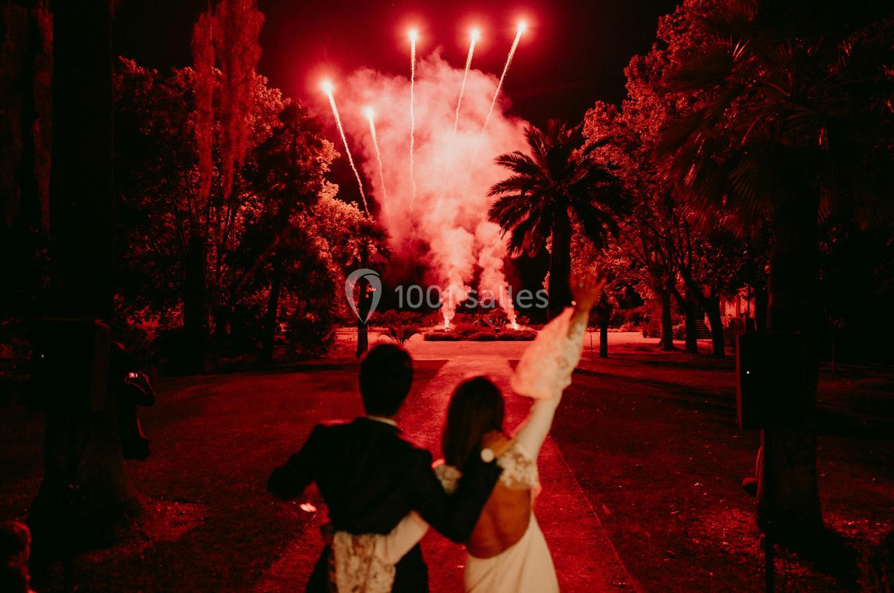 Location salle Le Cannet-des-Maures (Var) - Château Colbert Cannet #12 Un couple de dos regarde un feu d'artifice rouge éclatant dans un jardin la nuit.