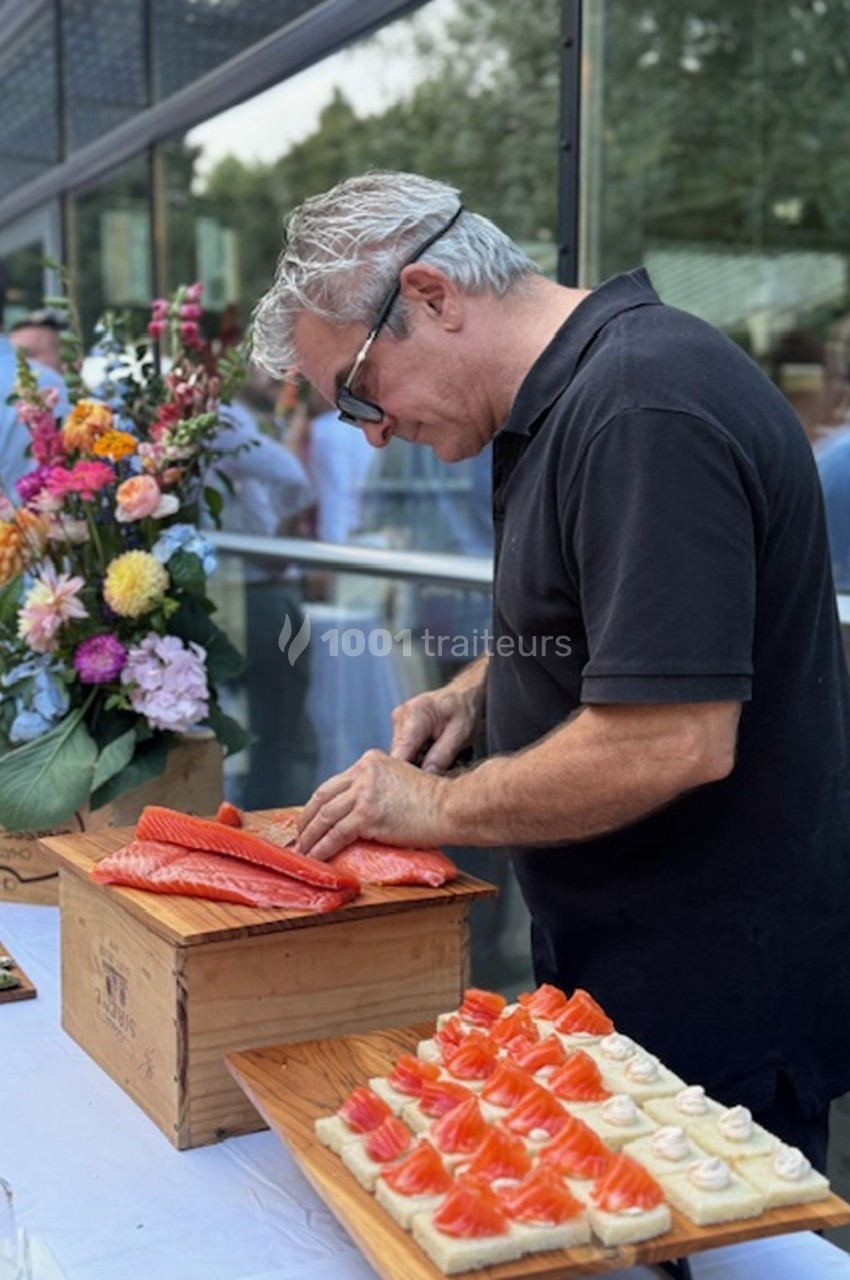 Un homme découpe du saumon frais sur une planche en bois, à côté de fleurs et de bouchées apéritives.