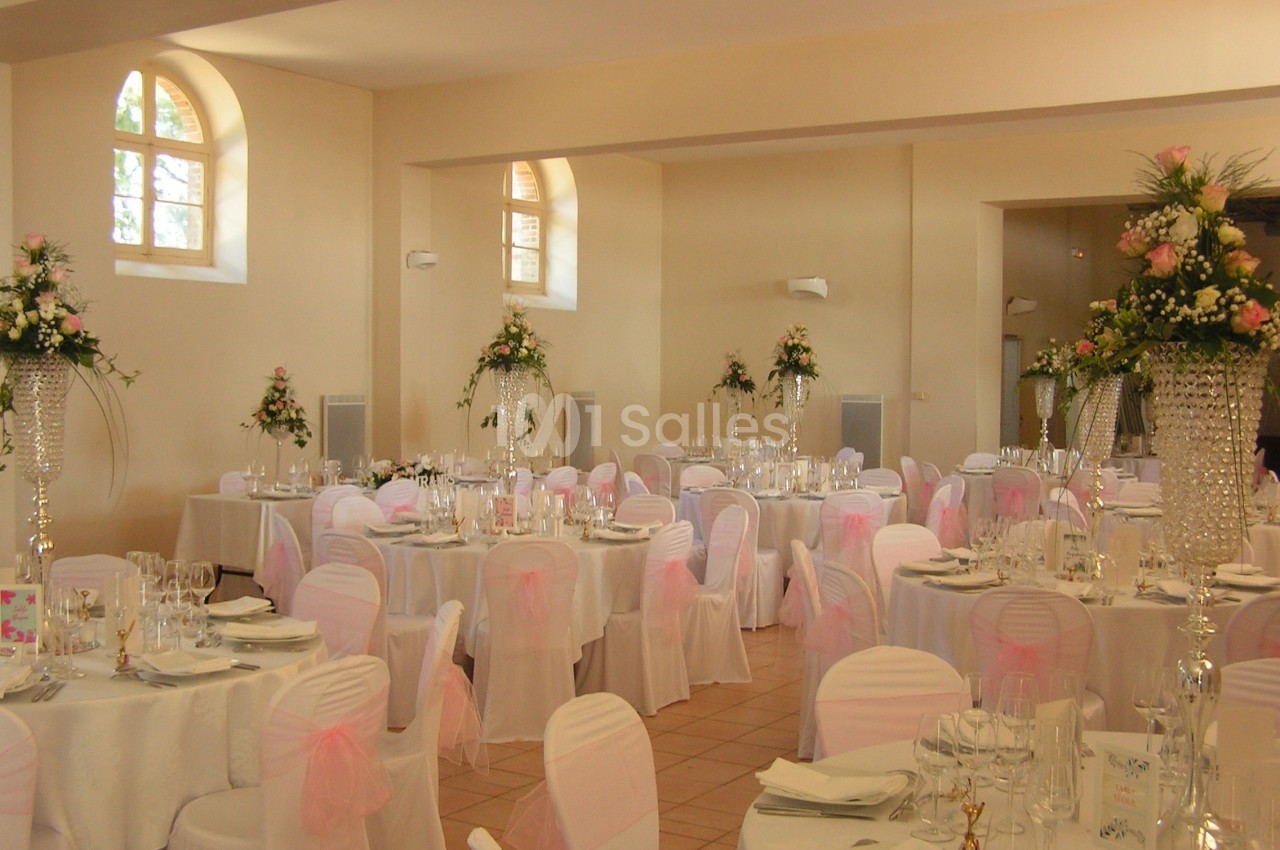 Salle de réception décorée avec des tables rondes, nappes blanches, chaises ornées de nœuds roses et compositions florales.