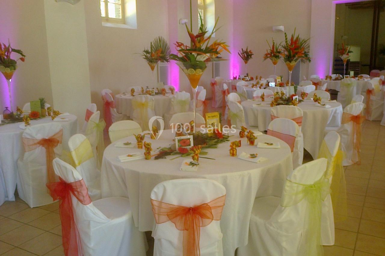 Salle décorée pour un événement avec des tables rondes, nappes blanches, chaises ornées de rubans colorés et centres de…