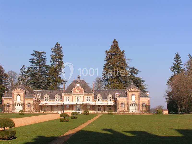Façade d'un manoir en briques entouré d'un grand jardin avec allée centrale, arbres et ciel dégagé en arrière-plan.