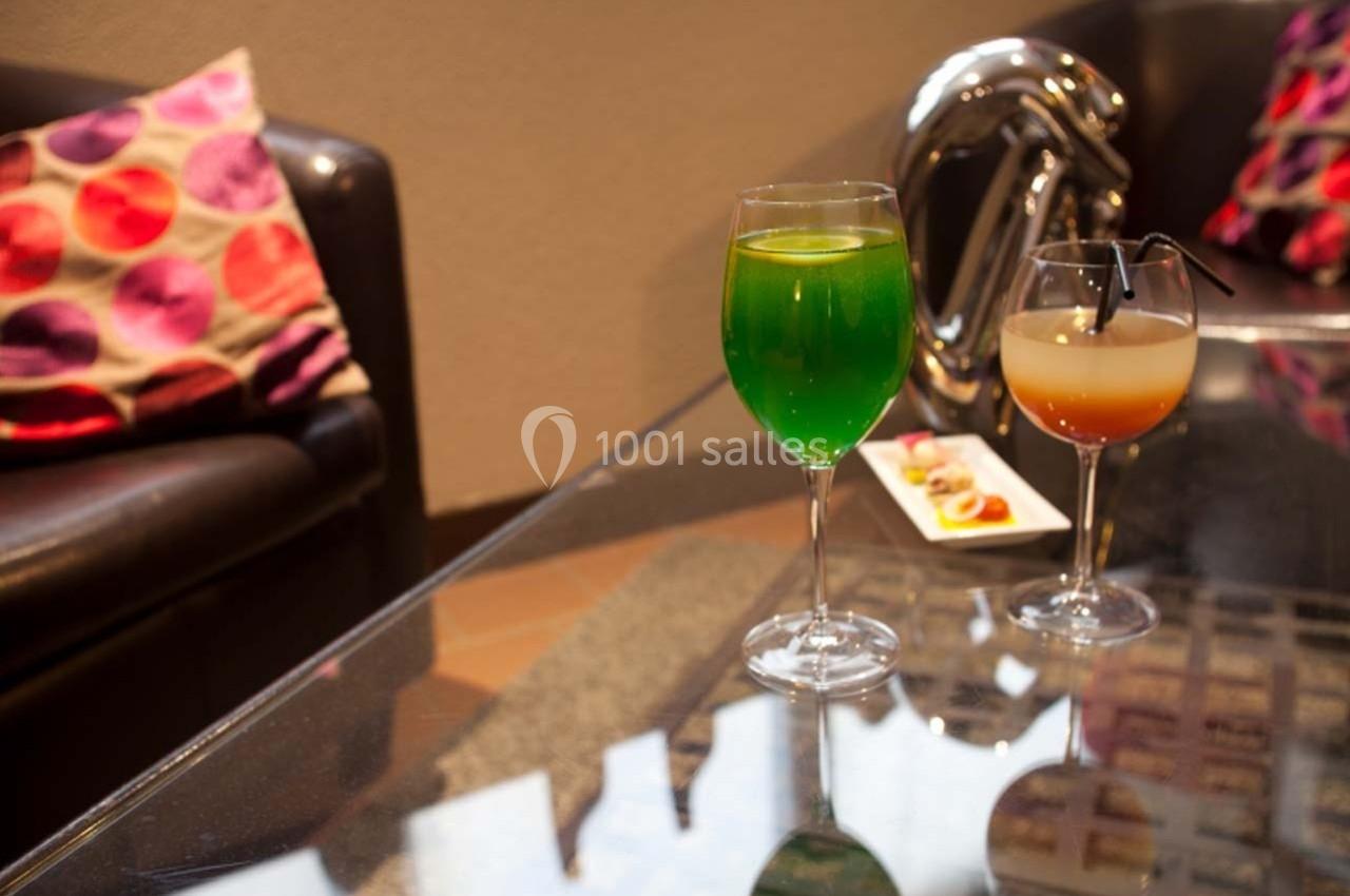 Location salle Bonneval (Eure-et-Loir) - Auberge de La Herse #5 Deux verres de cocktails colorés posés sur une table en verre, avec un canapé et des coussins en arrière-plan.