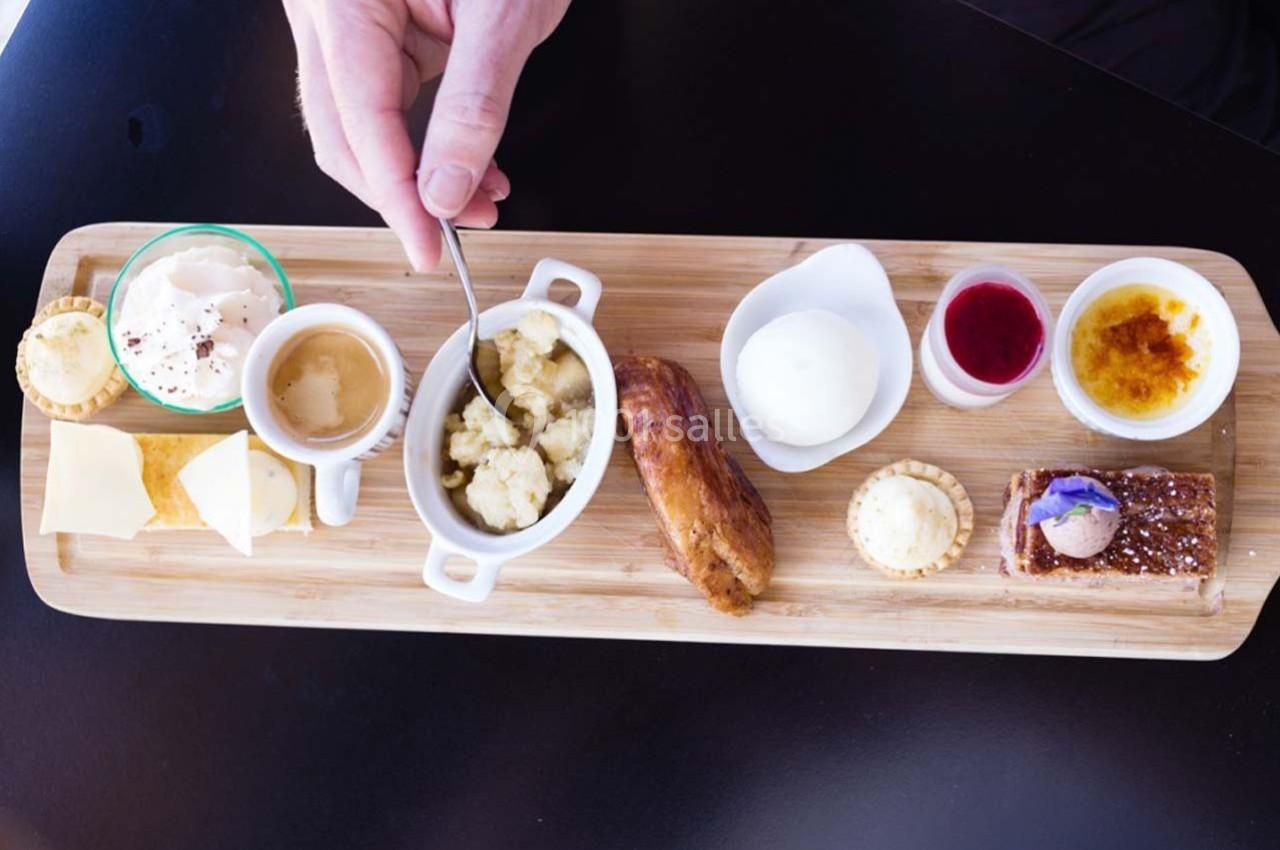 Location salle Bonneval (Eure-et-Loir) - Auberge de La Herse #14 Plateau en bois avec divers desserts, café, fromage, et condiments, une main tenant une cuillère au centre.