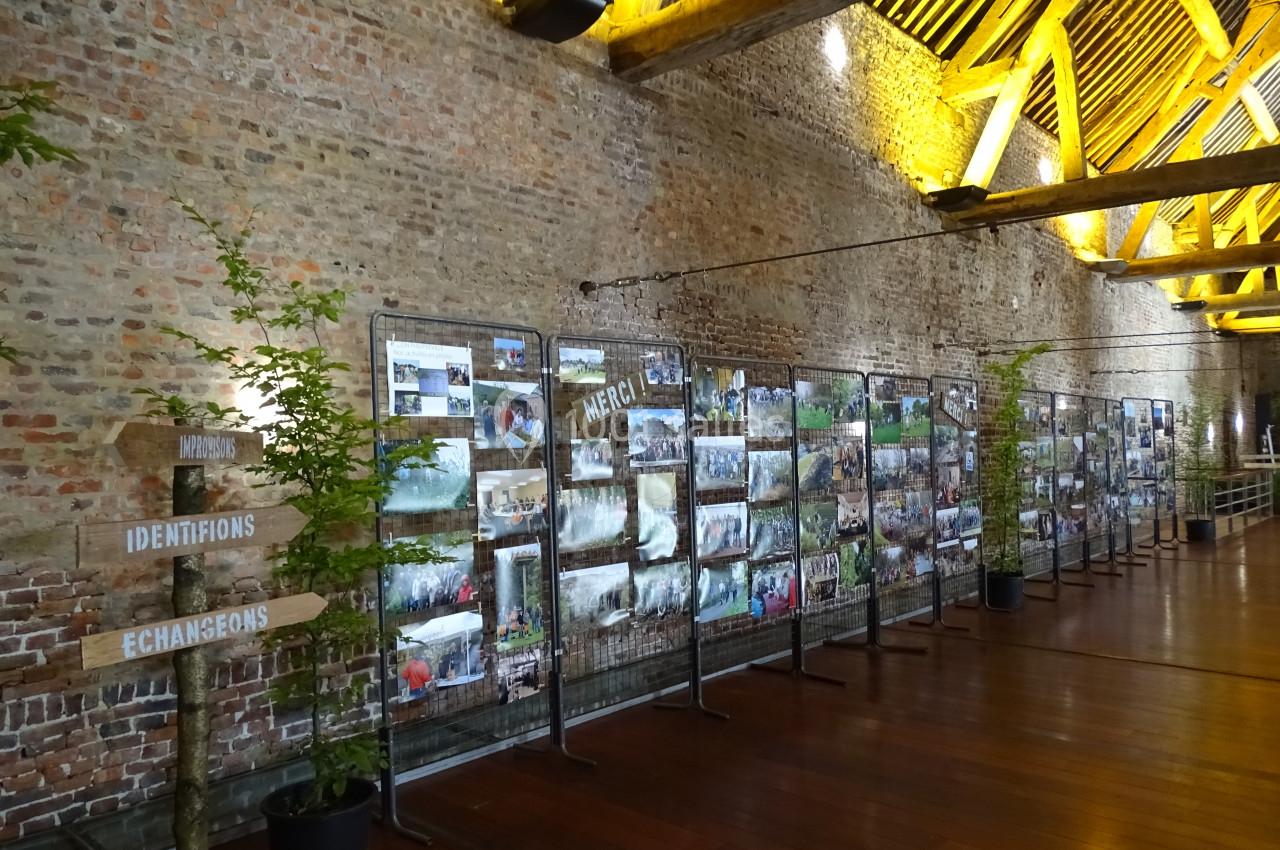 Location salle Anhée (Namur) - Ferme de l'Abbaye de Moulins #37 Exposition de photos accrochées sur des grilles dans une grande salle en briques avec un plafond en poutres apparentes.