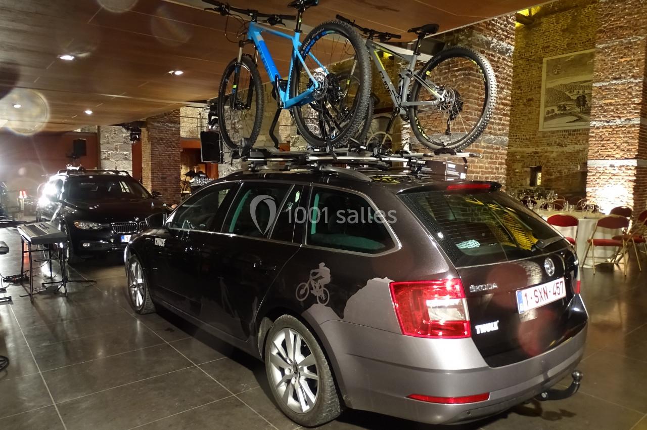 Location salle Anhée (Namur) - Ferme de l'Abbaye de Moulins #35 Voiture noire avec deux vélos fixés sur un porte-vélos de toit, stationnée dans un espace intérieur en briques.