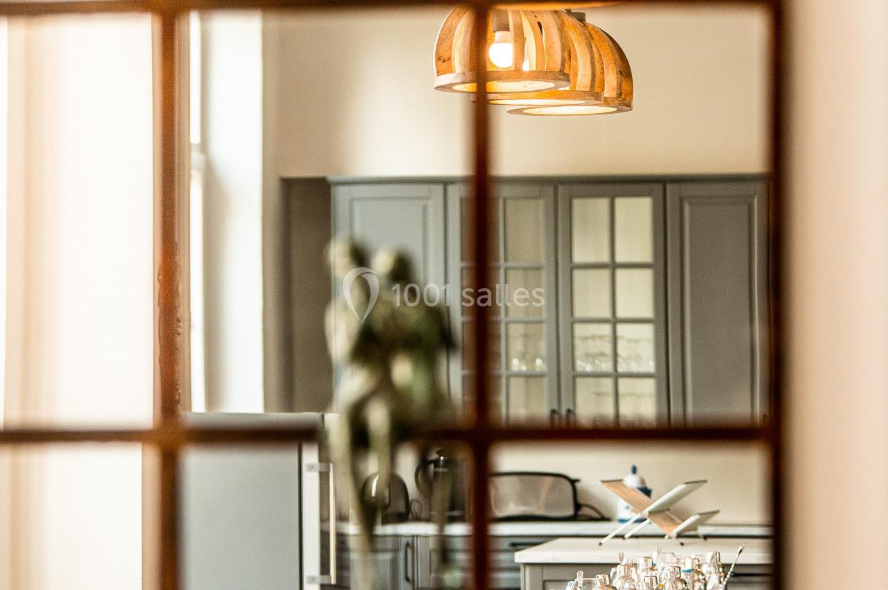 Location salle Anhée (Namur) - Ferme de l'Abbaye de Moulins #21 Vue d'une salle à manger élégante avec une table dressée, des chaises en osier et une cuisine en arrière-plan.
