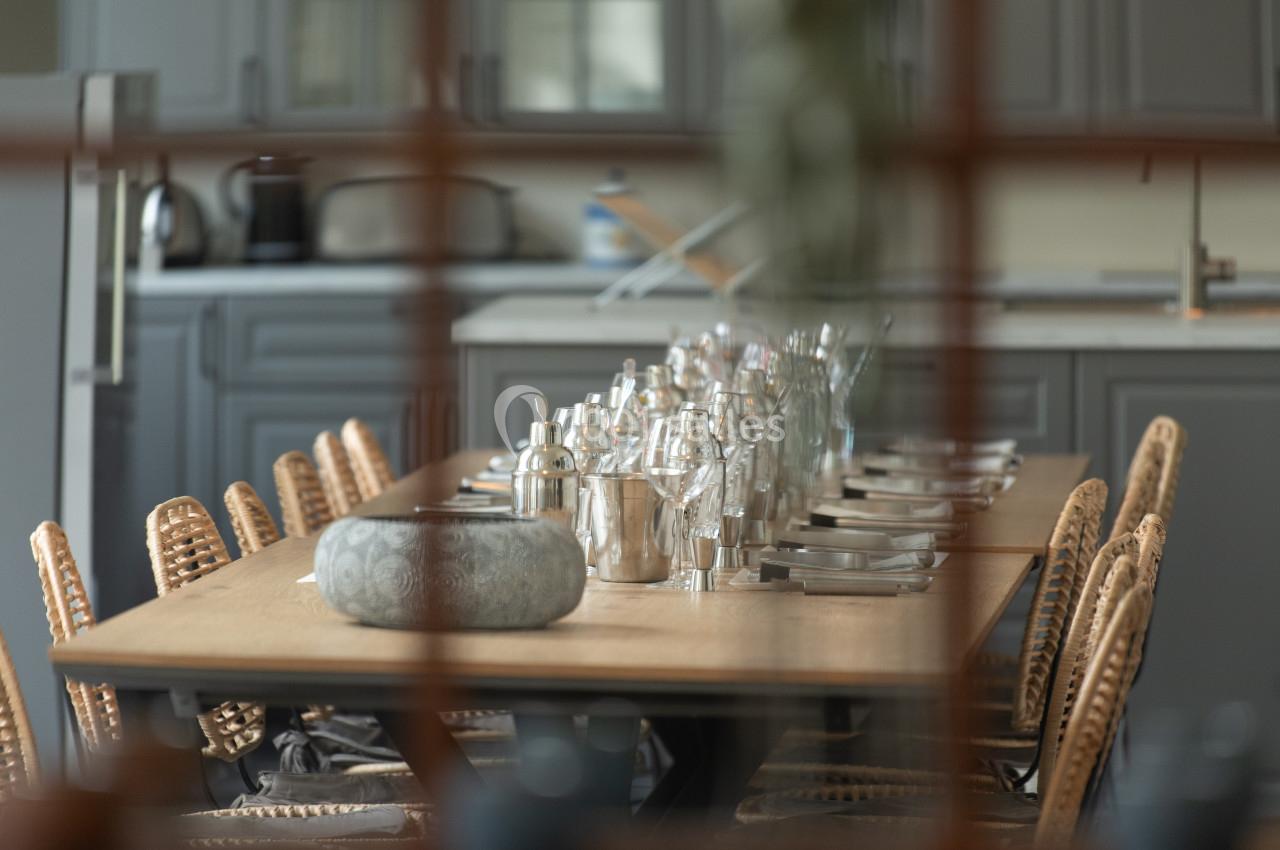 Location salle Anhée (Namur) - Ferme de l'Abbaye de Moulins #22 Table en bois dressée avec des couverts, des verres et des carafes, entourée de chaises en rotin dans une cuisine moderne.