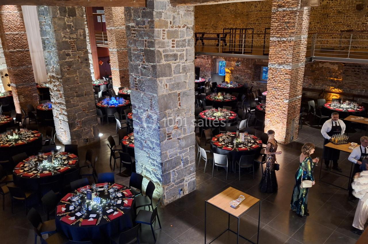 Location salle Anhée (Namur) - Ferme de l'Abbaye de Moulins #10 Salle en pierre avec tables rondes dressées pour un événement, éclairage tamisé et quelques personnes en tenue de soirée.