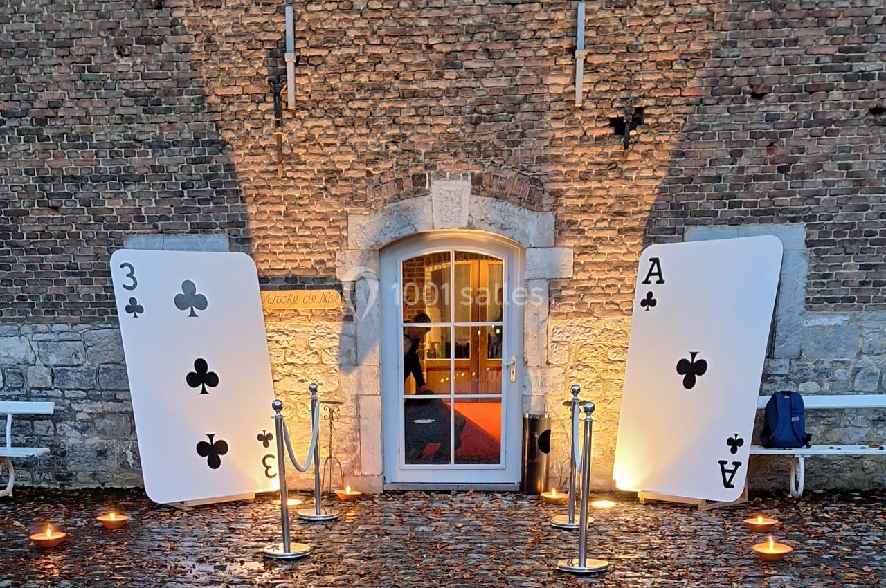 Location salle Anhée (Namur) - Ferme de l'Abbaye de Moulins #11 Deux grandes cartes à jouer (3 de trèfle et As de trèfle) encadrent une porte éclairée devant un mur en briques.