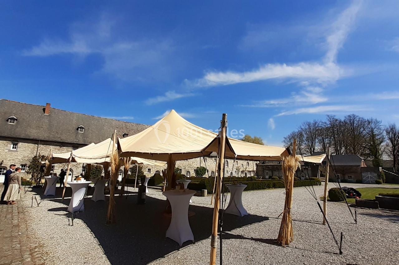 Location salle Anhée (Namur) - Ferme de l'Abbaye de Moulins #6 Tente beige installée dans une cour pavée, entourée de tables hautes et de bâtiments en pierre sous un ciel bleu.
