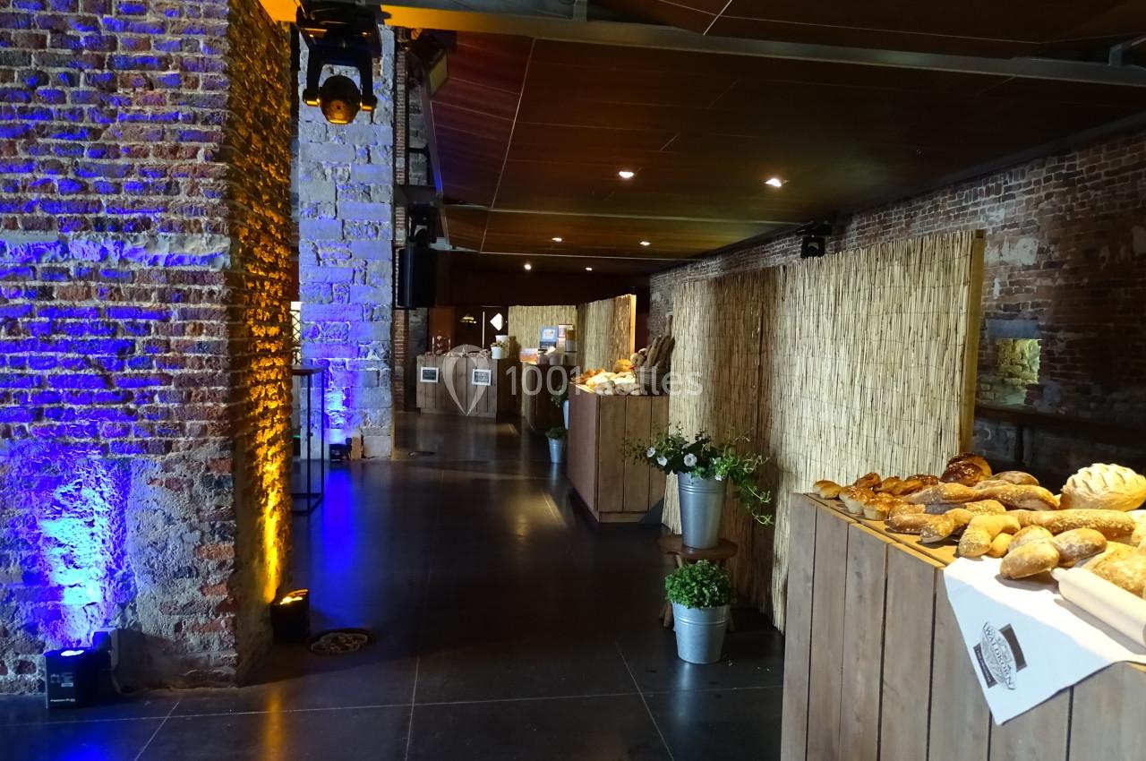 Location salle Anhée (Namur) - Ferme de l'Abbaye de Moulins #39 Salle avec murs en briques, éclairage tamisé, tables en bois garnies de pains et décorations végétales.