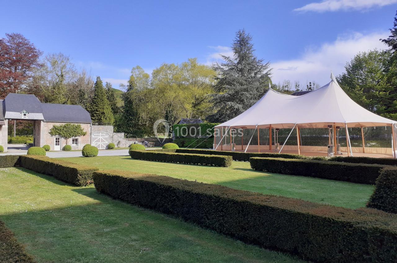 Location salle Anhée (Namur) - Ferme de l'Abbaye de Moulins #4 Jardin avec pelouse, haies taillées, tente blanche et bâtiment en pierre sous un ciel bleu.