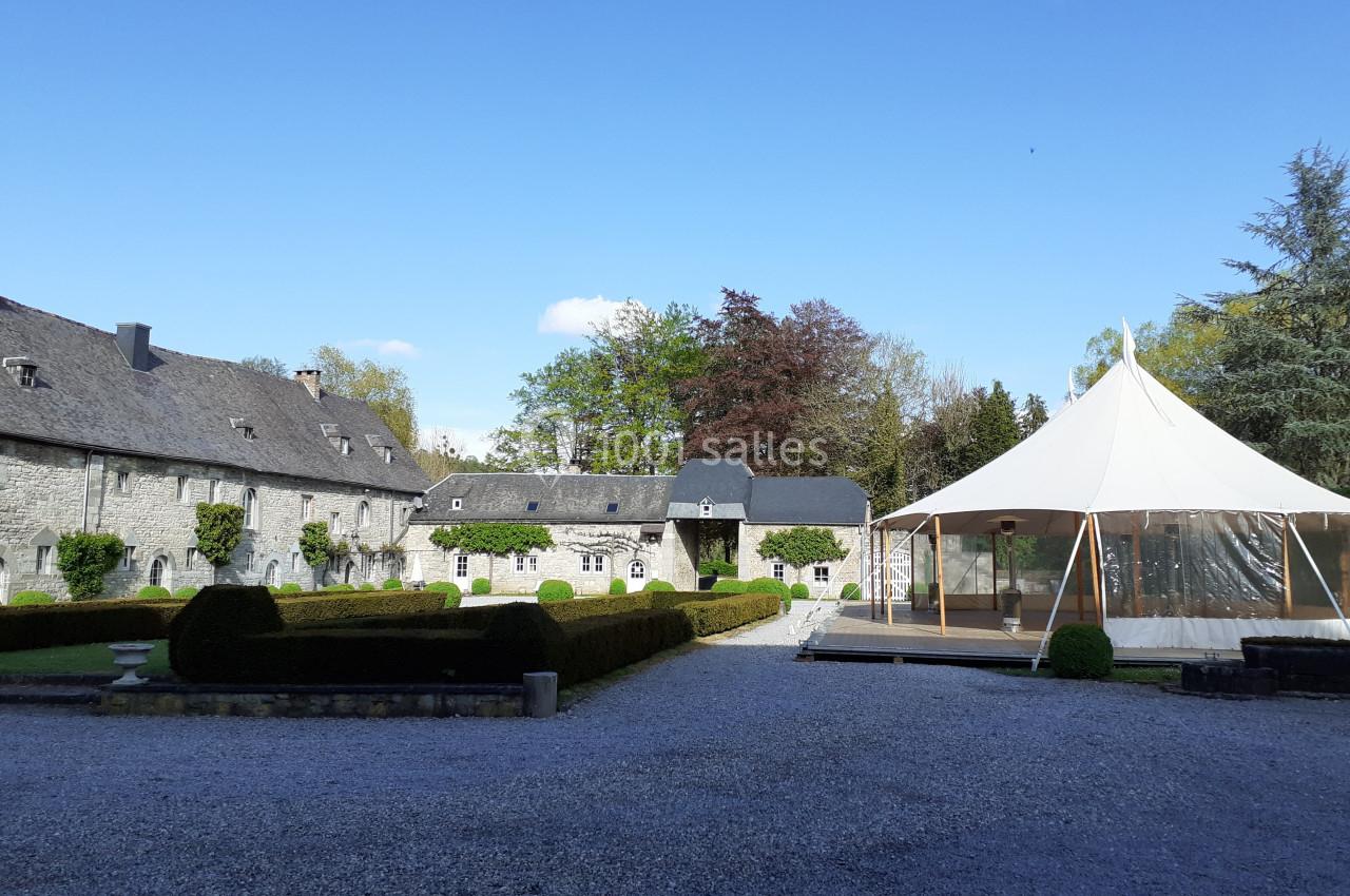 Location salle Anhée (Namur) - Ferme de l'Abbaye de Moulins #13 Cour d’un domaine avec bâtiments en pierre, haies taillées et une tente blanche sur une terrasse en bois.