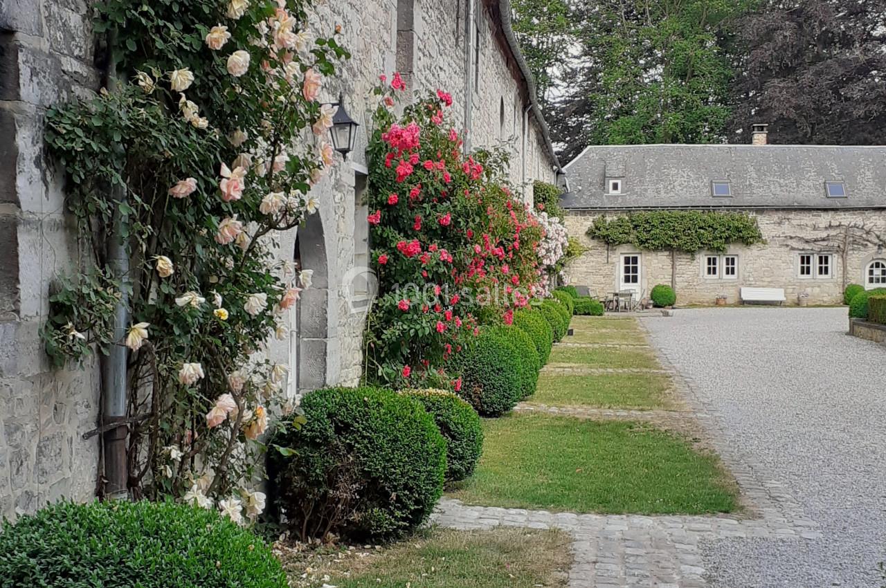 Location salle Anhée (Namur) - Ferme de l'Abbaye de Moulins #23 Façade en pierre d'un bâtiment ancien avec des rosiers grimpants, allée pavée et jardin soigné.