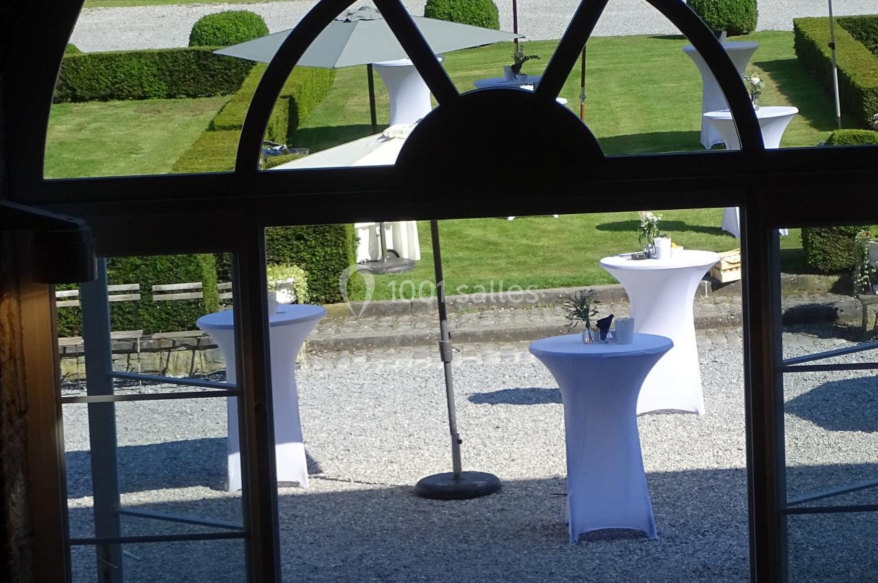 Location salle Anhée (Namur) - Ferme de l'Abbaye de Moulins #26 Vue depuis une porte vitrée sur une cour pavée avec des tables hautes dressées et un jardin en arrière-plan.