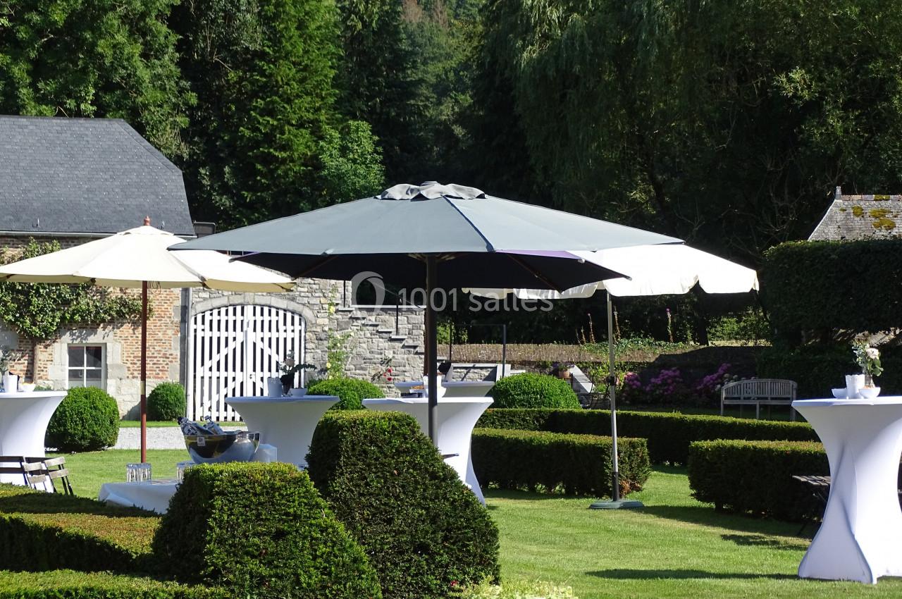 Location salle Anhée (Namur) - Ferme de l'Abbaye de Moulins #19 Parasols blancs et gris installés dans un jardin verdoyant avec des tables hautes et des haies taillées.