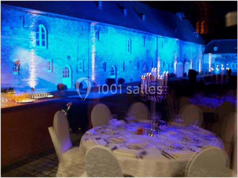 Location salle Anhée (Namur) - Ferme de l'Abbaye de Moulins #28 Salle de réception éclairée en bleu avec une table dressée au premier plan et un bâtiment en pierre en arrière-plan.